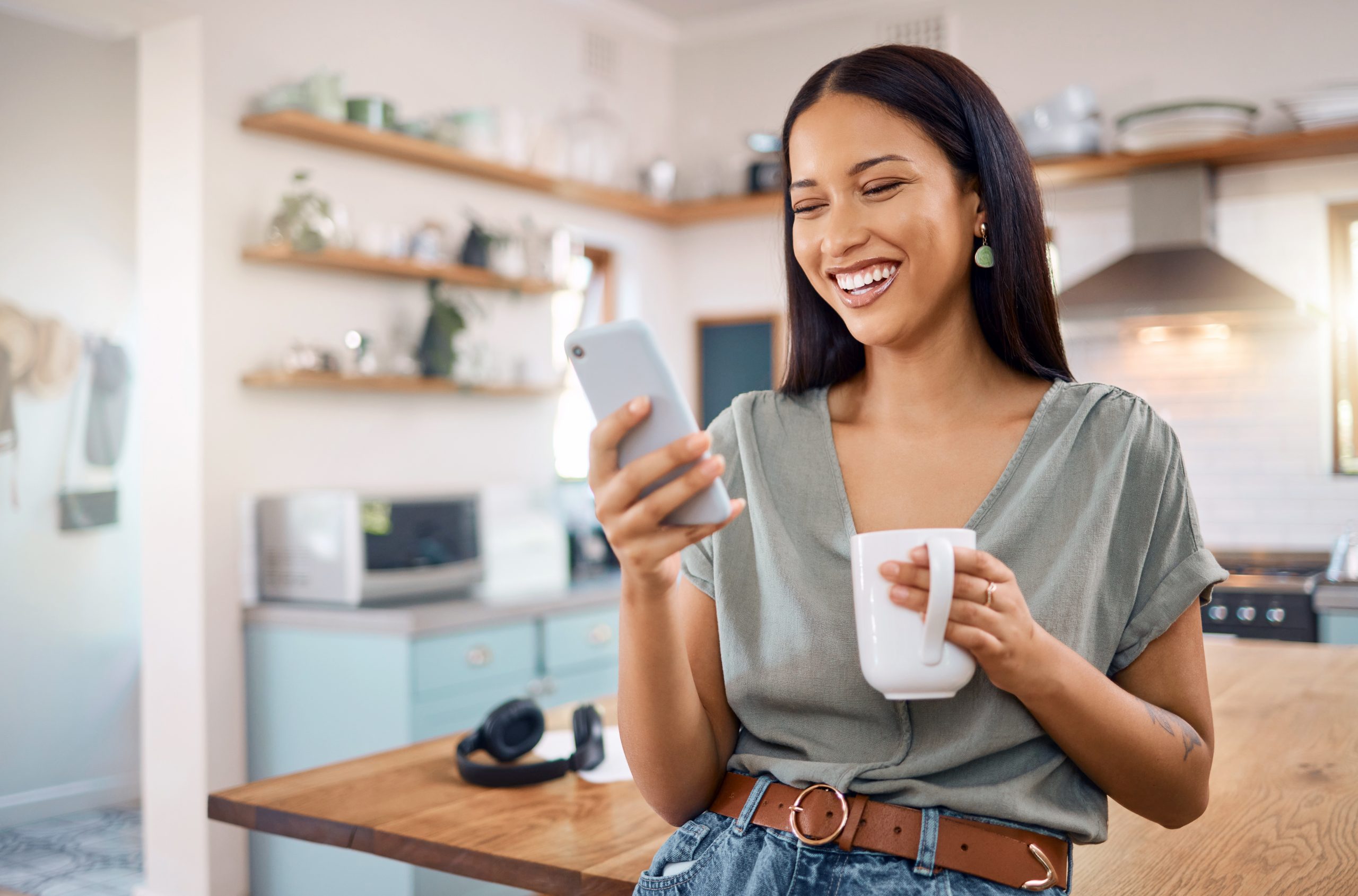 One happy young mixed race woman standing in her kitchen at home and using smartphone to browse the internet while drinking a cup of coffee. Smiling hispanic on social media and networking on a phone - Spotless gutters